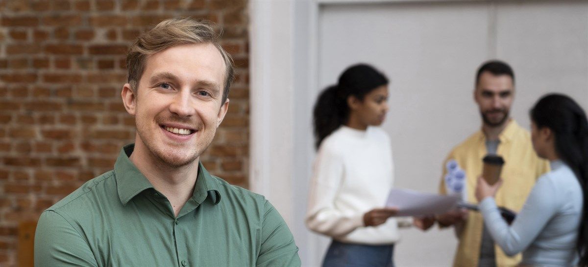 A man looking into the camera and smiling, while a group of 3 professional converse in the background.