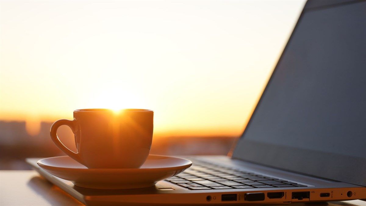 A tea cup and saucer sitting on top of a laptop. The sunset can be seen in the background peaking over the cup